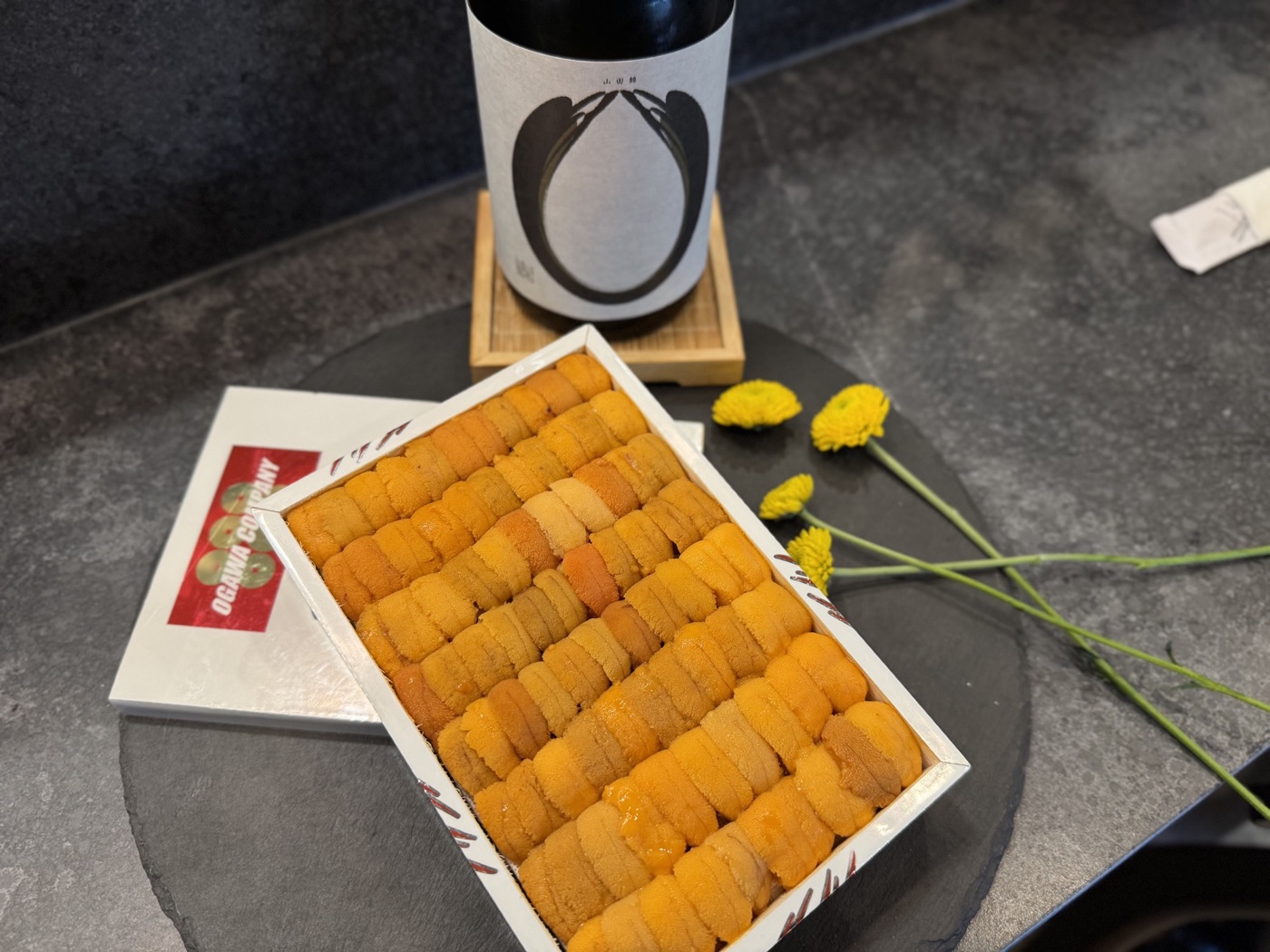 An angled view of a sea urchin tray with a sake bottle and yellow flowers